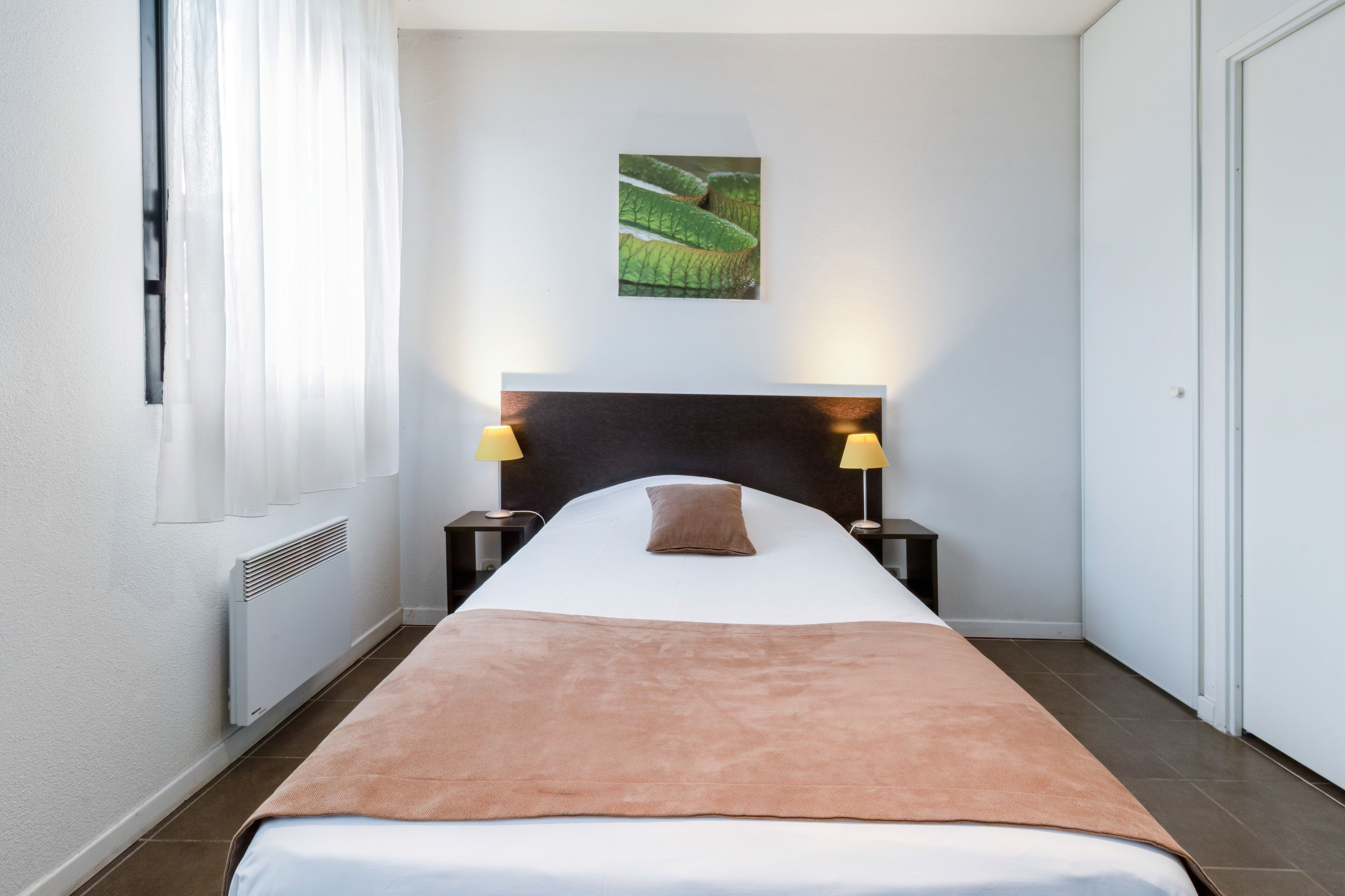 Habitación de hotel con cama, dos lámparas de noche, cabecera, radiador, ventana con cortinas e imagen colgada en la pared.