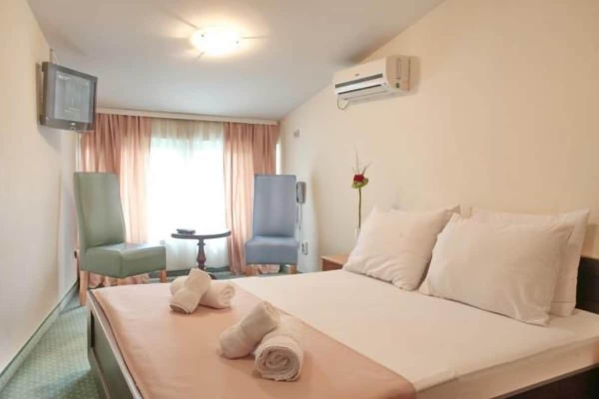 A hotel room with a bed, two chairs, a small table, an air conditioner, and a TV.
