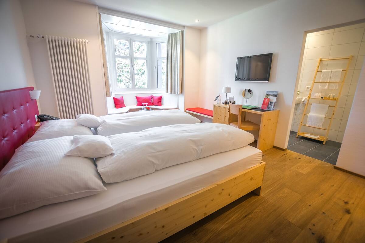 A hotel room with two beds, a desk, a TV, and a bathroom with a wooden ladder.