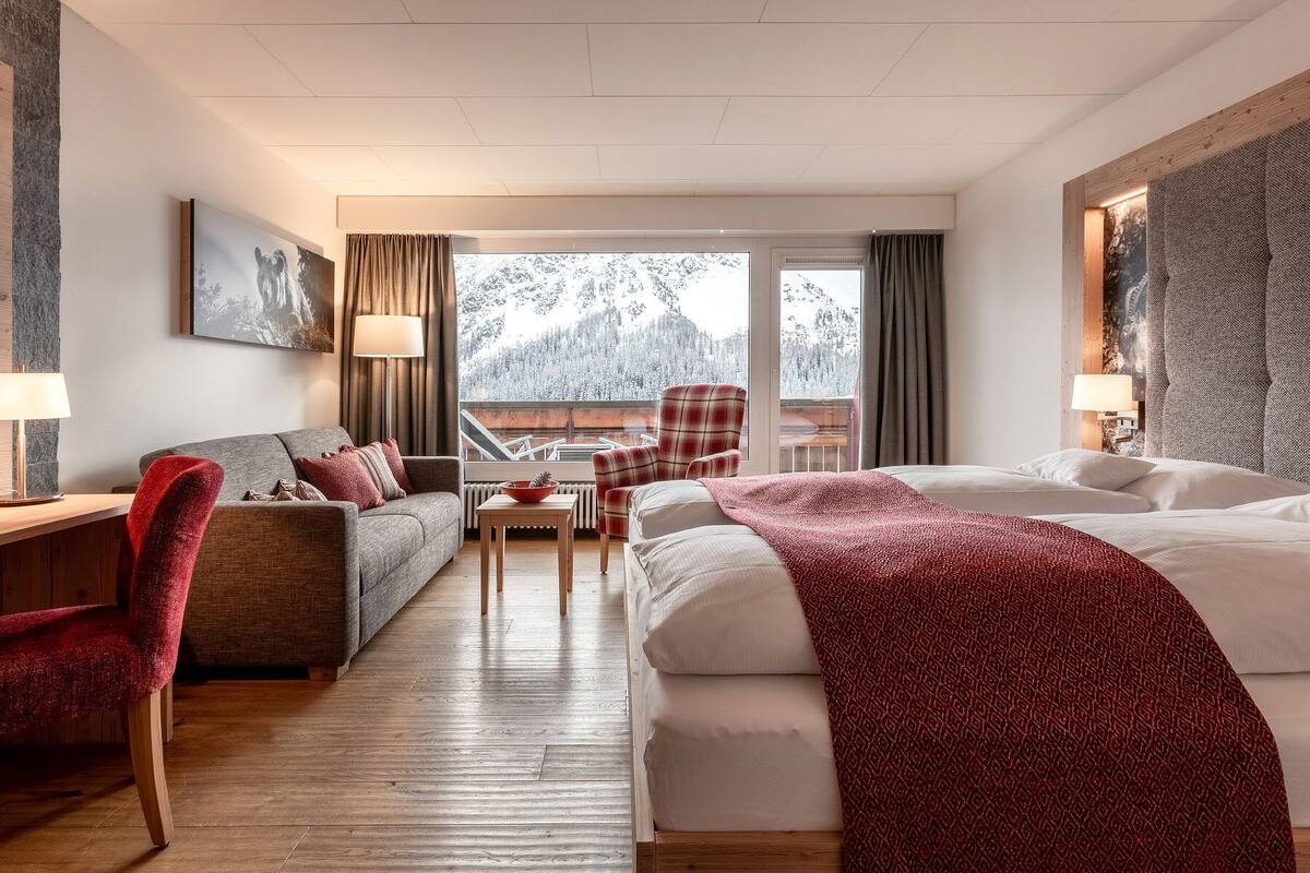 A hotel room with a large bed, a sofa, a chair, a table, and a view of snow-covered trees through the window.