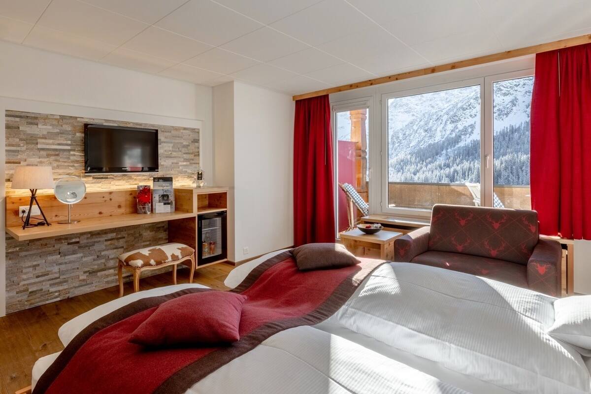 A hotel room with a bed, a desk with a TV, a small table, and a chair. There is a window with a view of snow-covered mountains.