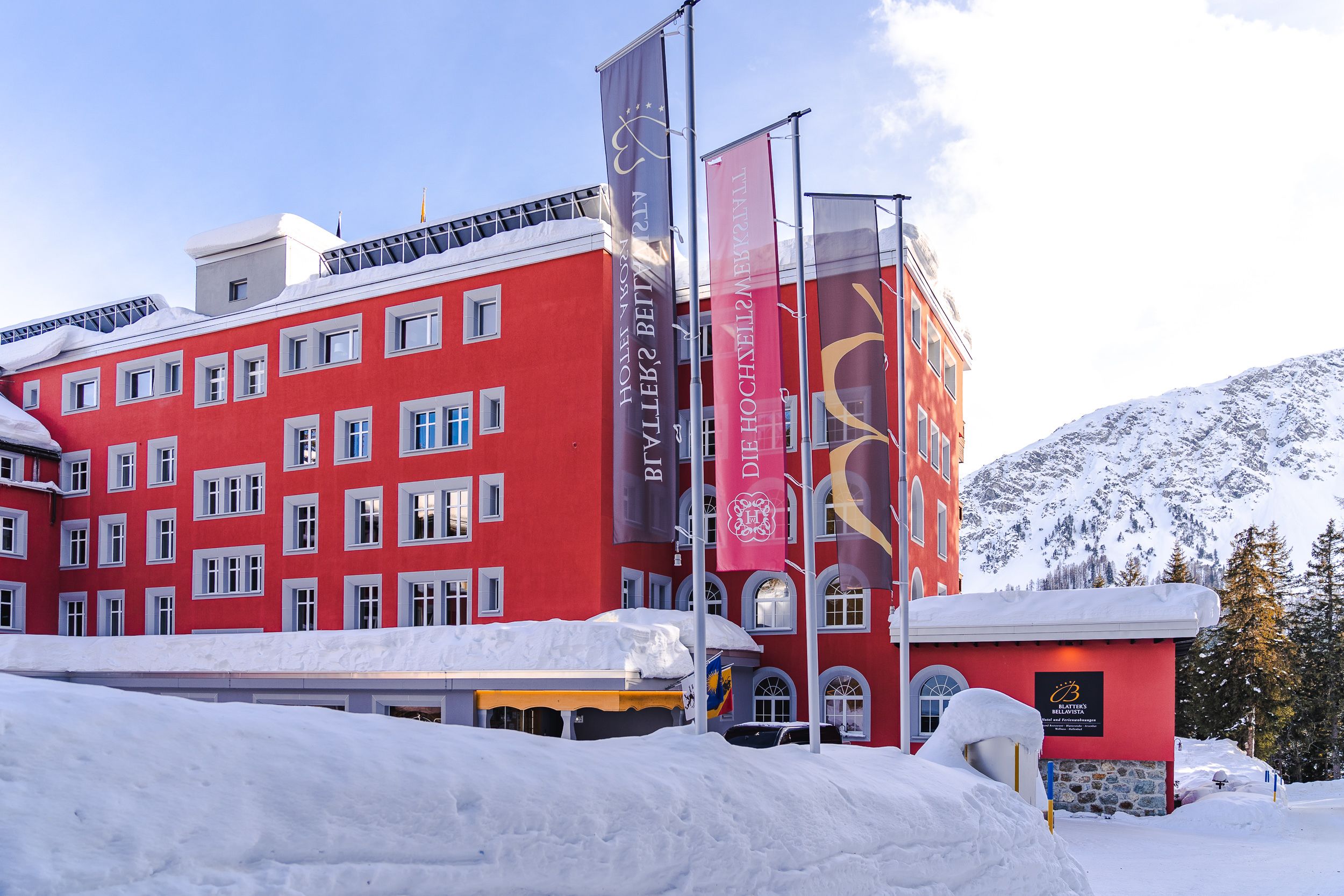 A red building with multiple windows and flags, surrounded by snow and mountains.