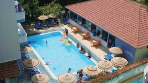 Une piscine extérieure, dotée de chaises longues et de parasols, est entourée d’un bâtiment dont le toit est recouvert de tuiles.