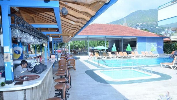 Un bar au bord de la piscine, doté de nombreux tabourets et d’un barman, une piscine avec des chaises longues, et une vue sur les montagnes en arrière-plan.