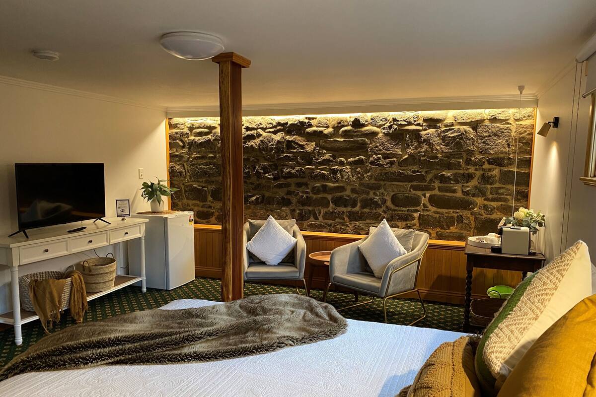 A hotel room with a stone wall, a bed with a fur throw, a TV on a white dresser, and two chairs with cushions.