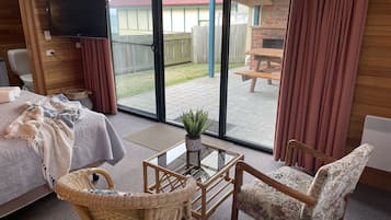 A room with a bed, a wicker chair, a glass table, and a view of an outdoor patio with a table and chairs.