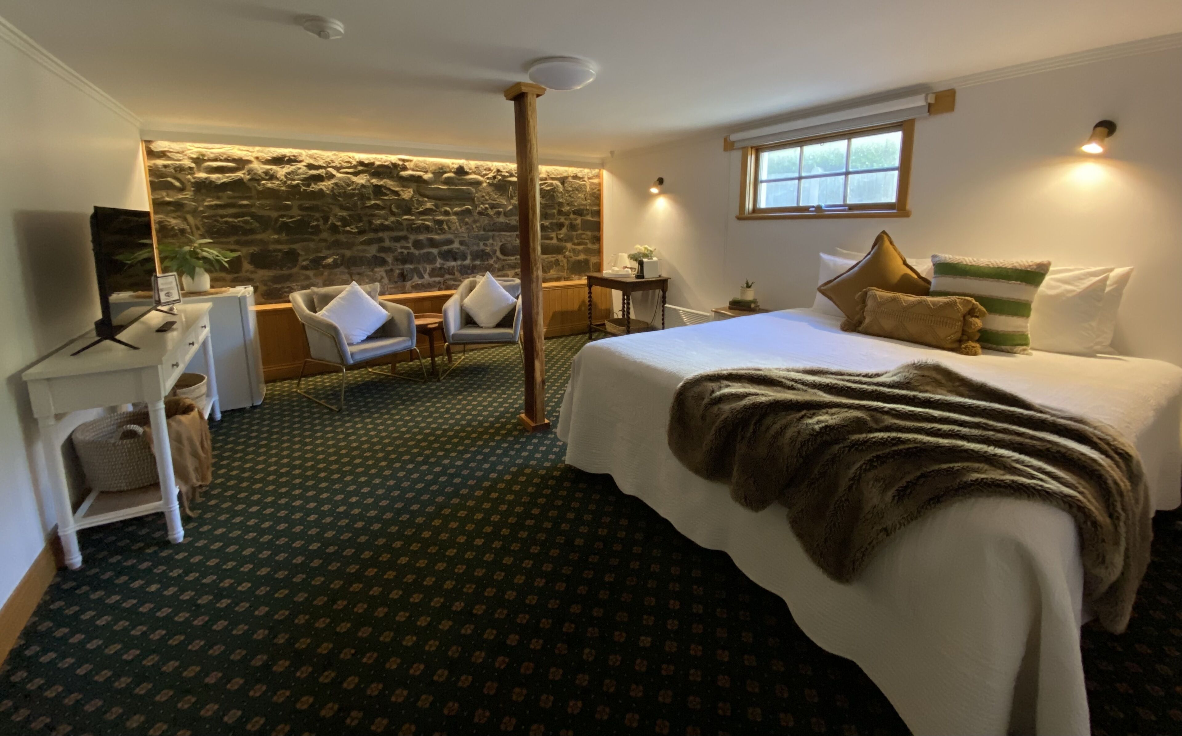 A hotel room with a large bed, a stone wall, a small table, a chair, and a television.