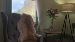 A room with a tufted armchair, a vase with flowers, a lamp, and a view of a mountain through a window. - Hanlon Guest House (Stanley)
