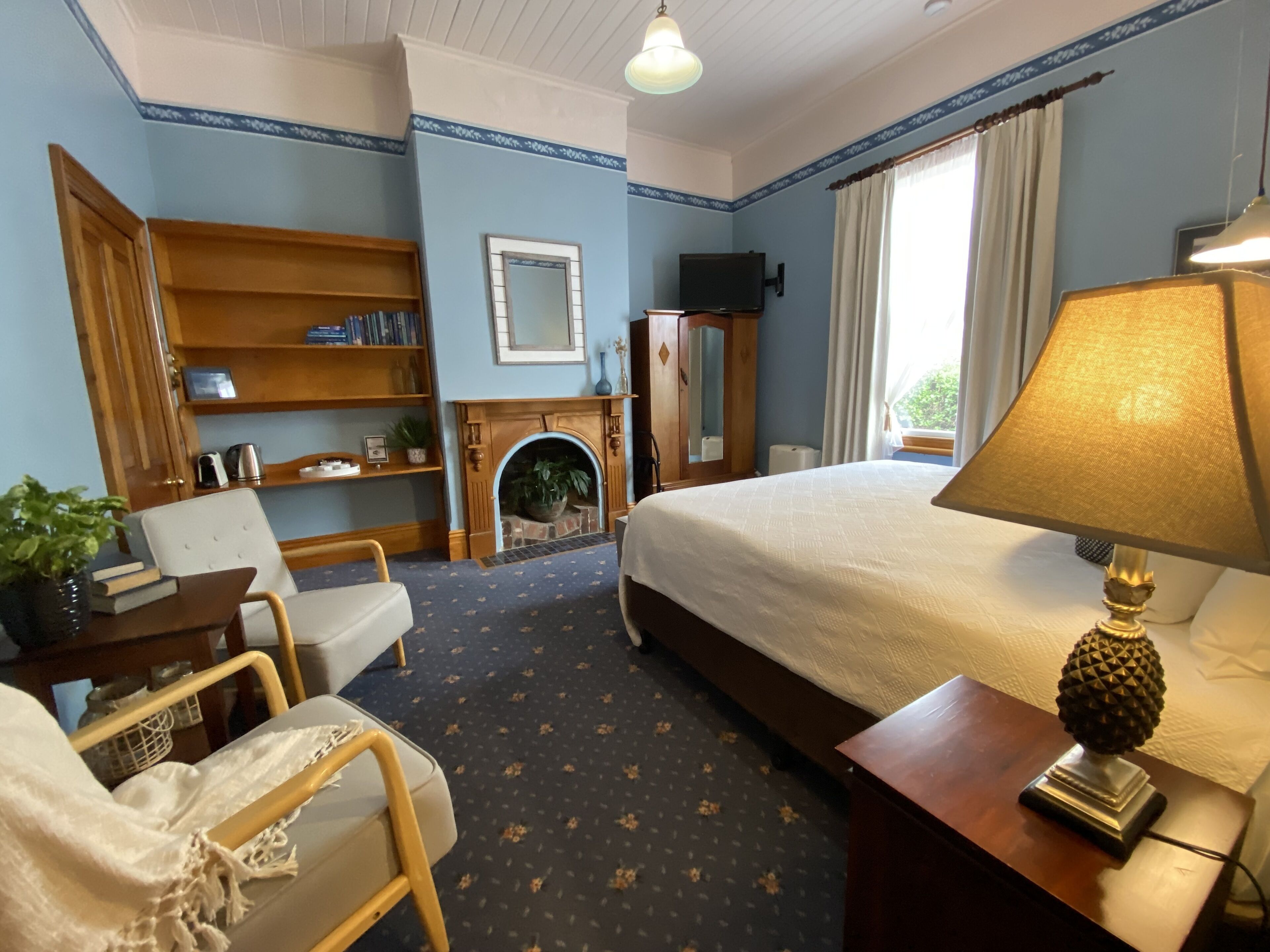 A hotel room with a bed, a desk with a lamp, a chair, a bookshelf, a fireplace, and a window with curtains.
