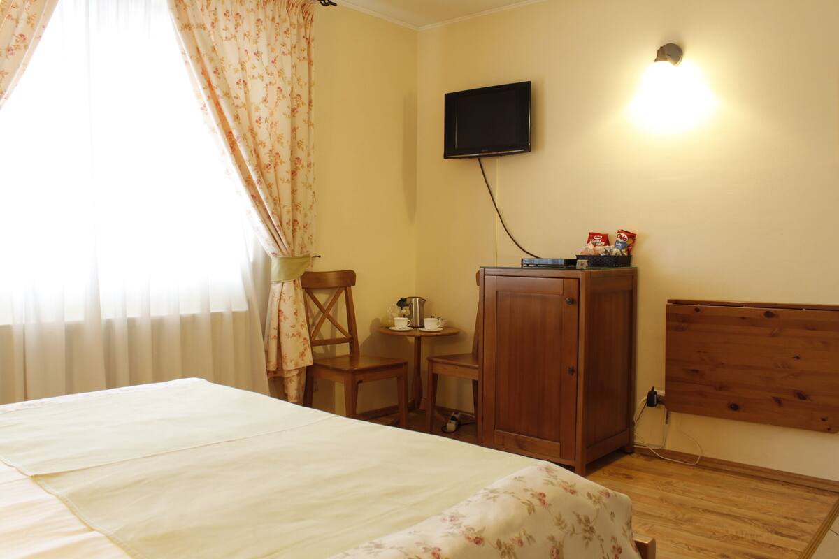 Quarto de hotel com uma cama, um armário de madeira, uma televisão na parede, uma mesinha com chávenas e uma chaleira, e uma cadeira.