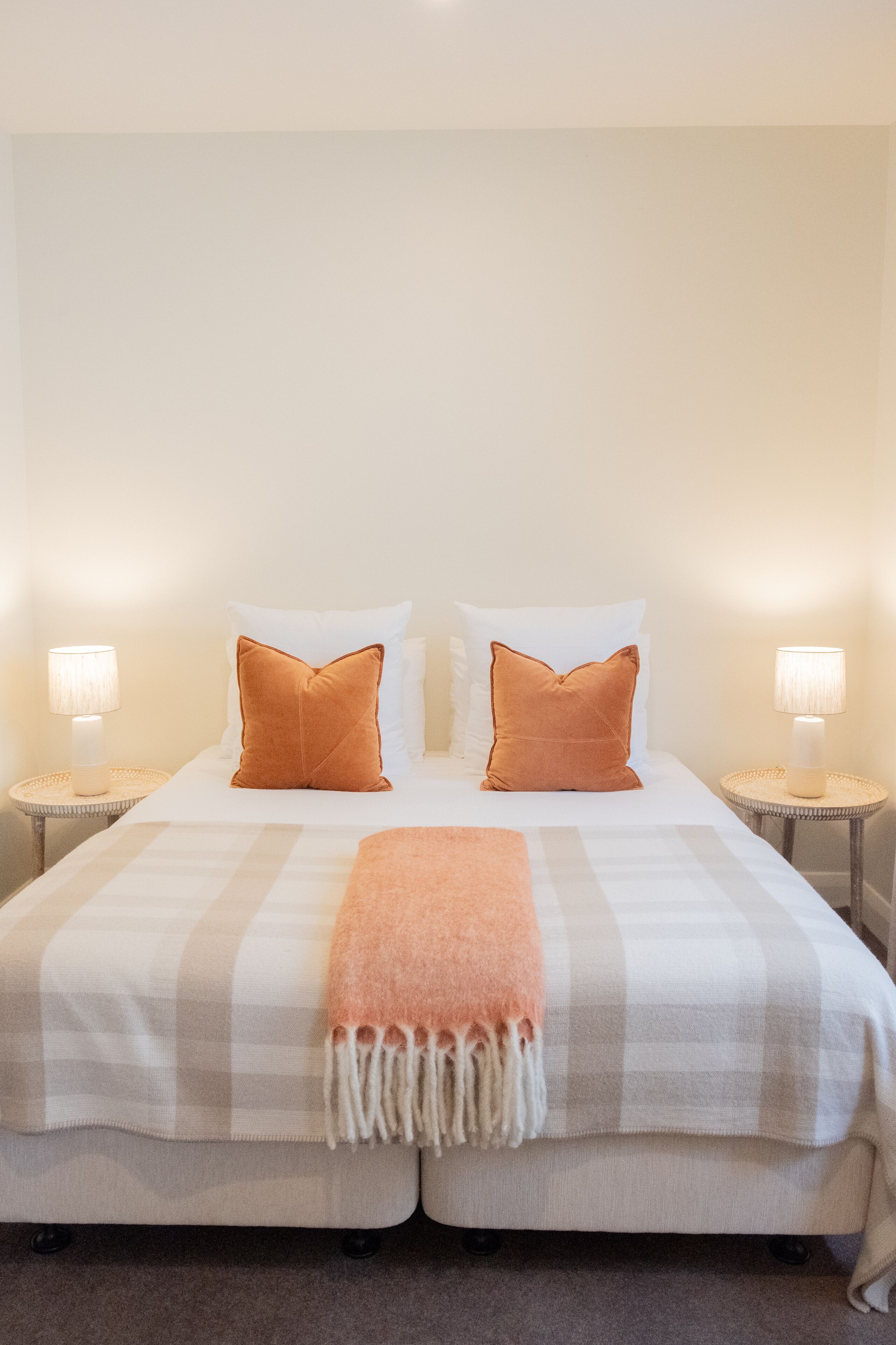 Una cama doble con dos almohadas blancas y dos naranjas, una colcha a cuadros y dos lámparas de noche sobre mesitas redondas.