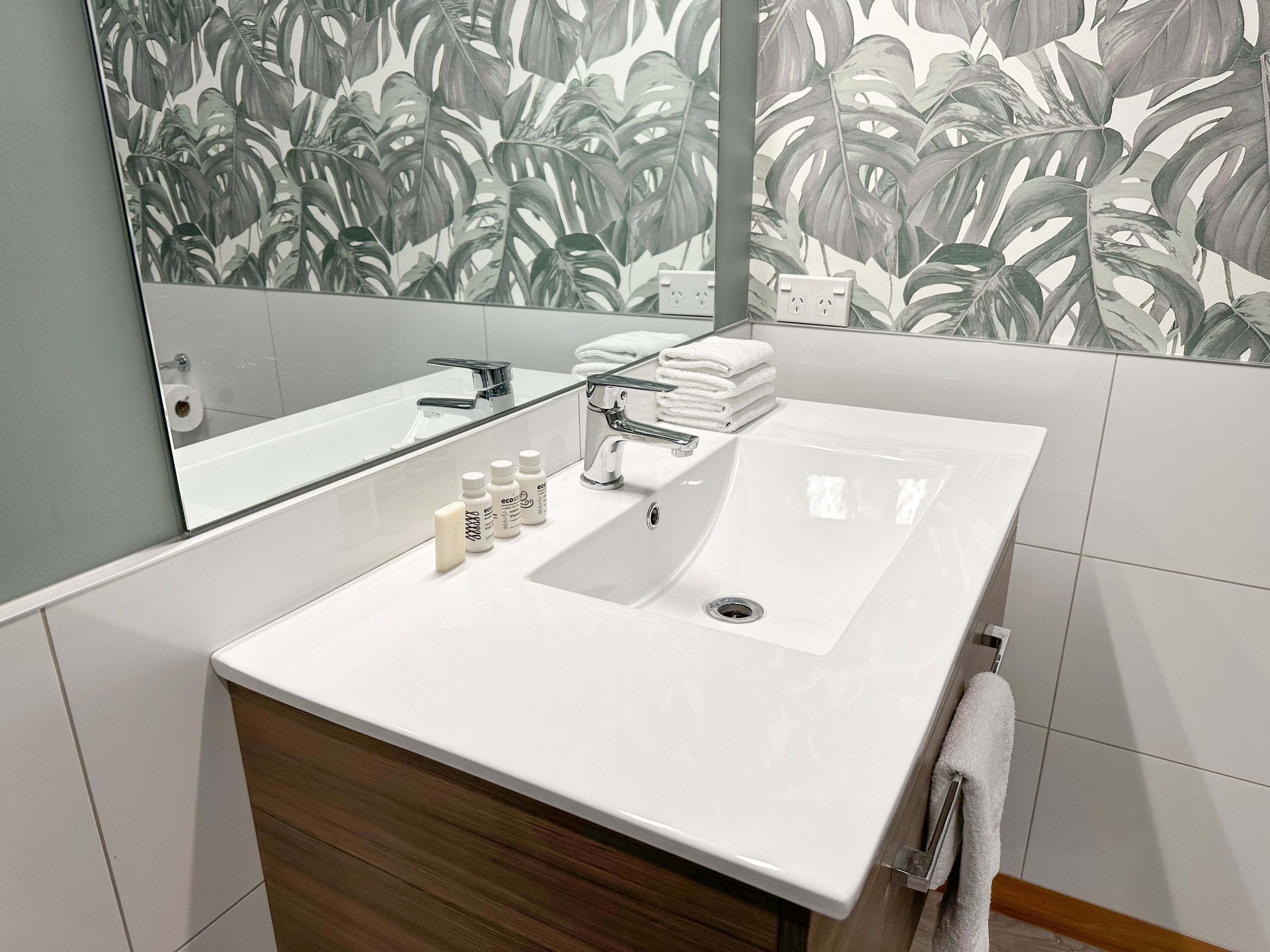 Ein modernes Badezimmer mit Waschbecken in Weiß, einem Spiegel und tropischer Blattmuster-Tapete.