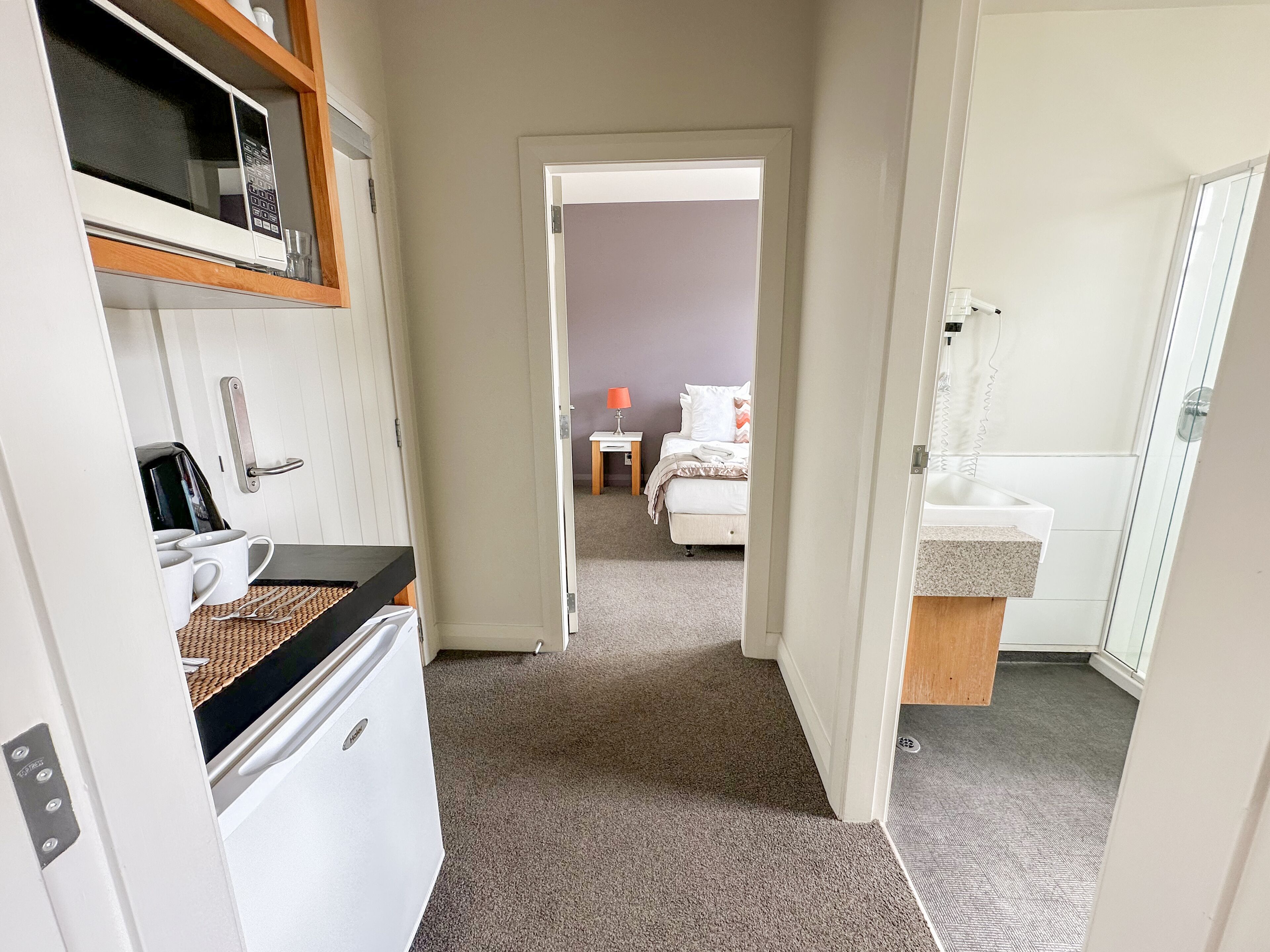 A narrow hallway leading to a bedroom with a bed, a bedside table, and a lamp. A bathroom with a sink and mirror is visible to the right.