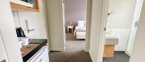 A narrow hallway leading to a bedroom with a bed, a bedside table, and a lamp. A bathroom with a sink and mirror is visible to the right.