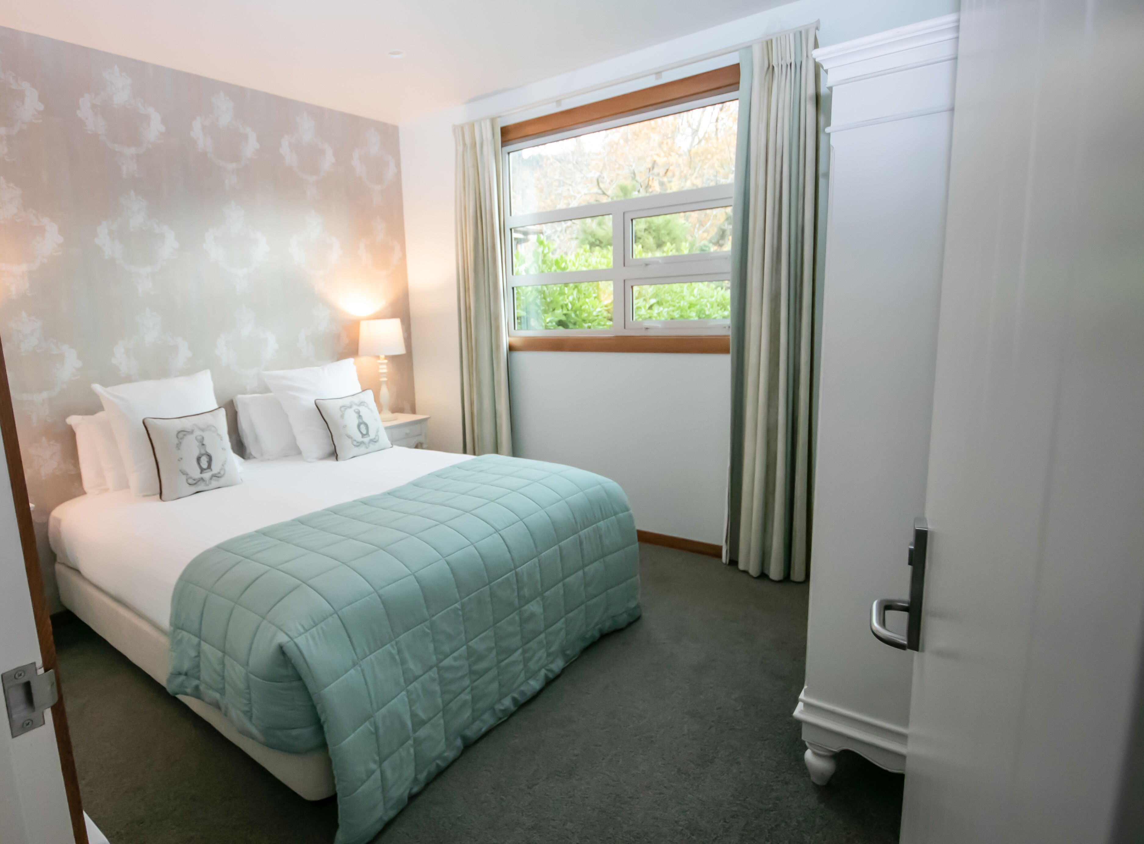 A neatly made bed with a quilted cover, a bedside table with a lamp, and a window with curtains.