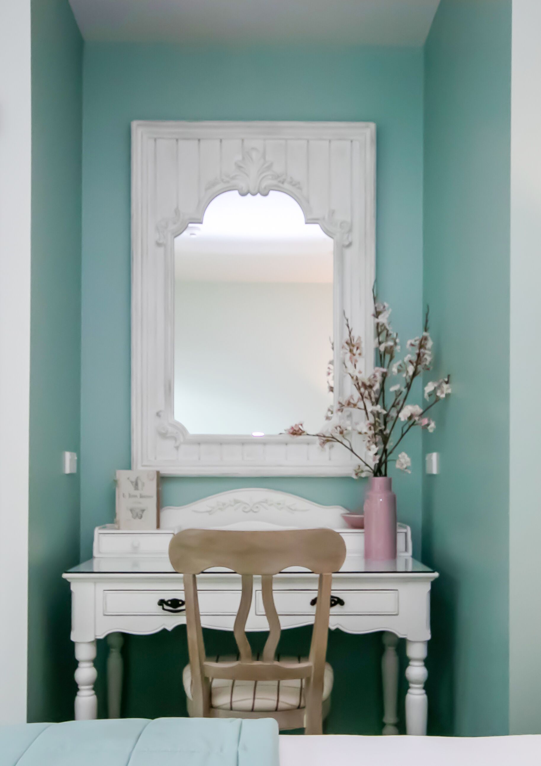 Una habitación con paredes color turquesa, un tocador blanco con espejo, una silla de madera y un jarrón rosa con ramas.