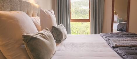 Una cama bien hecha con cabecera, una ventana con cortinas y vista a follaje exterior.