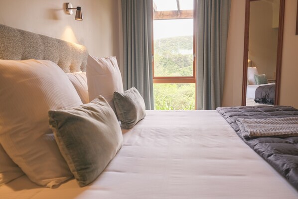 A neatly made bed with a headboard, a window with curtains, and a view of greenery outside.