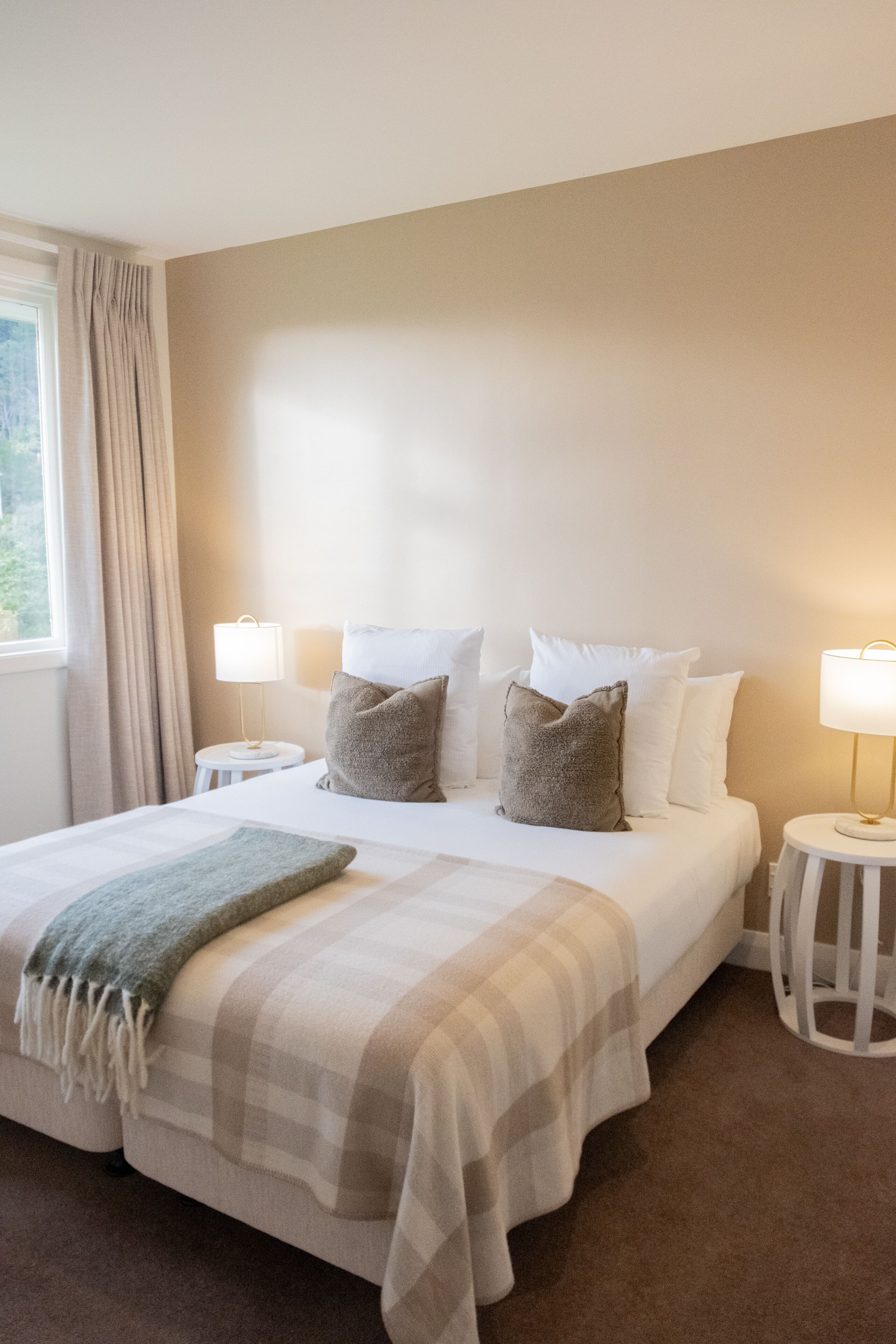 Een opgemaakt bed met een geruit plaid, twee bedlampjes en een raam met gordijnen.