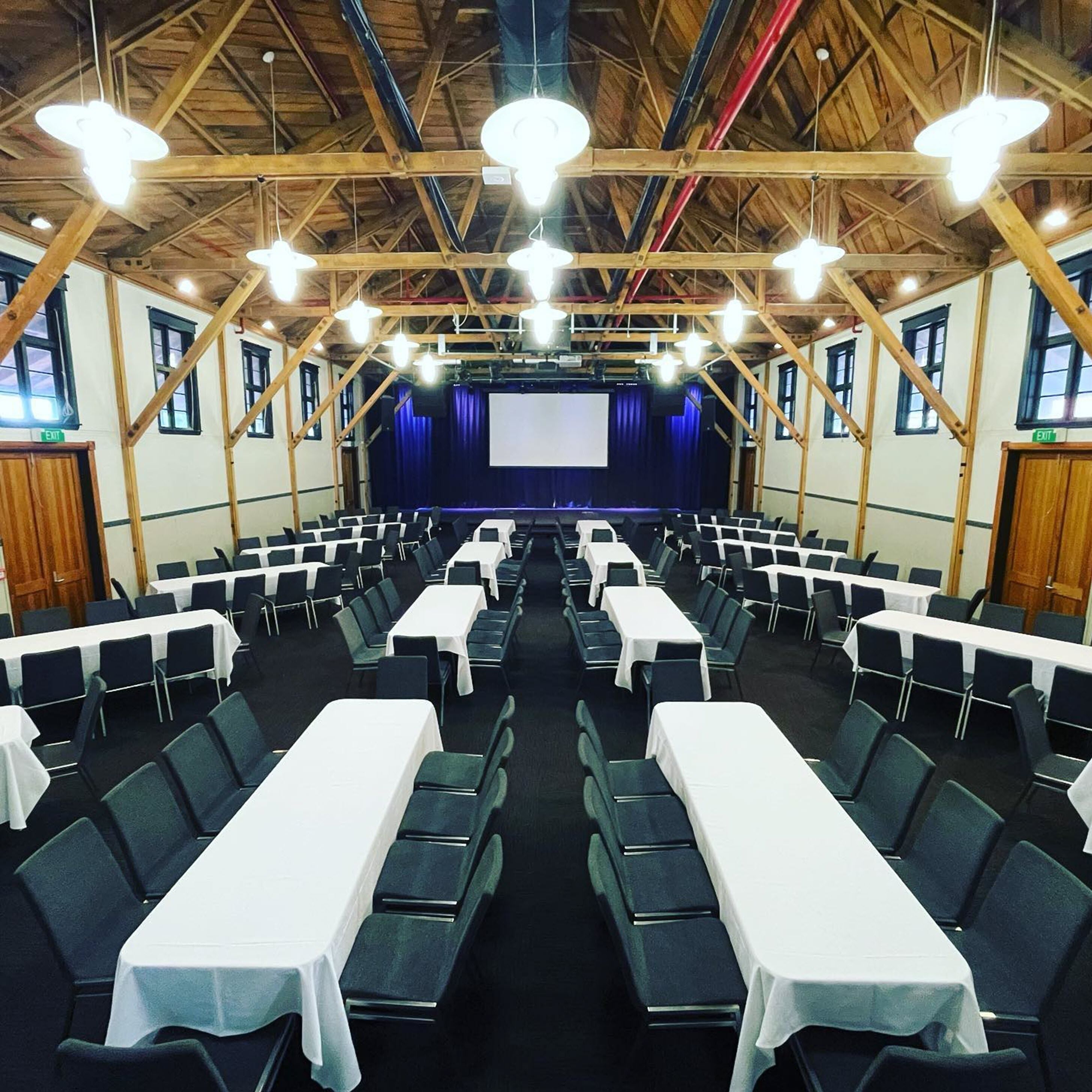 Een congrescentrum met rijen tafels en stoelen, een podium met een scherm en houten balken aan het plafond.