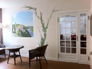 Una habitación con un cuadro de paisaje, una puerta con paneles de vidrio, muebles de mimbre y una lámpara.