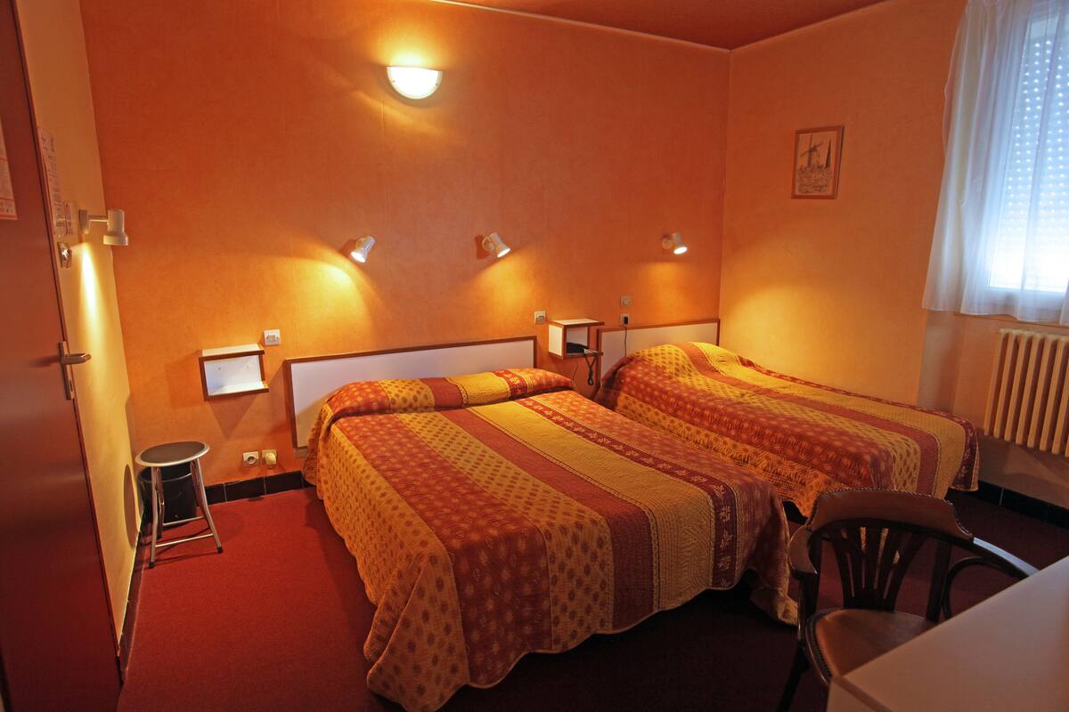 A hotel room with two beds, a small stool, a chair, and a desk.
