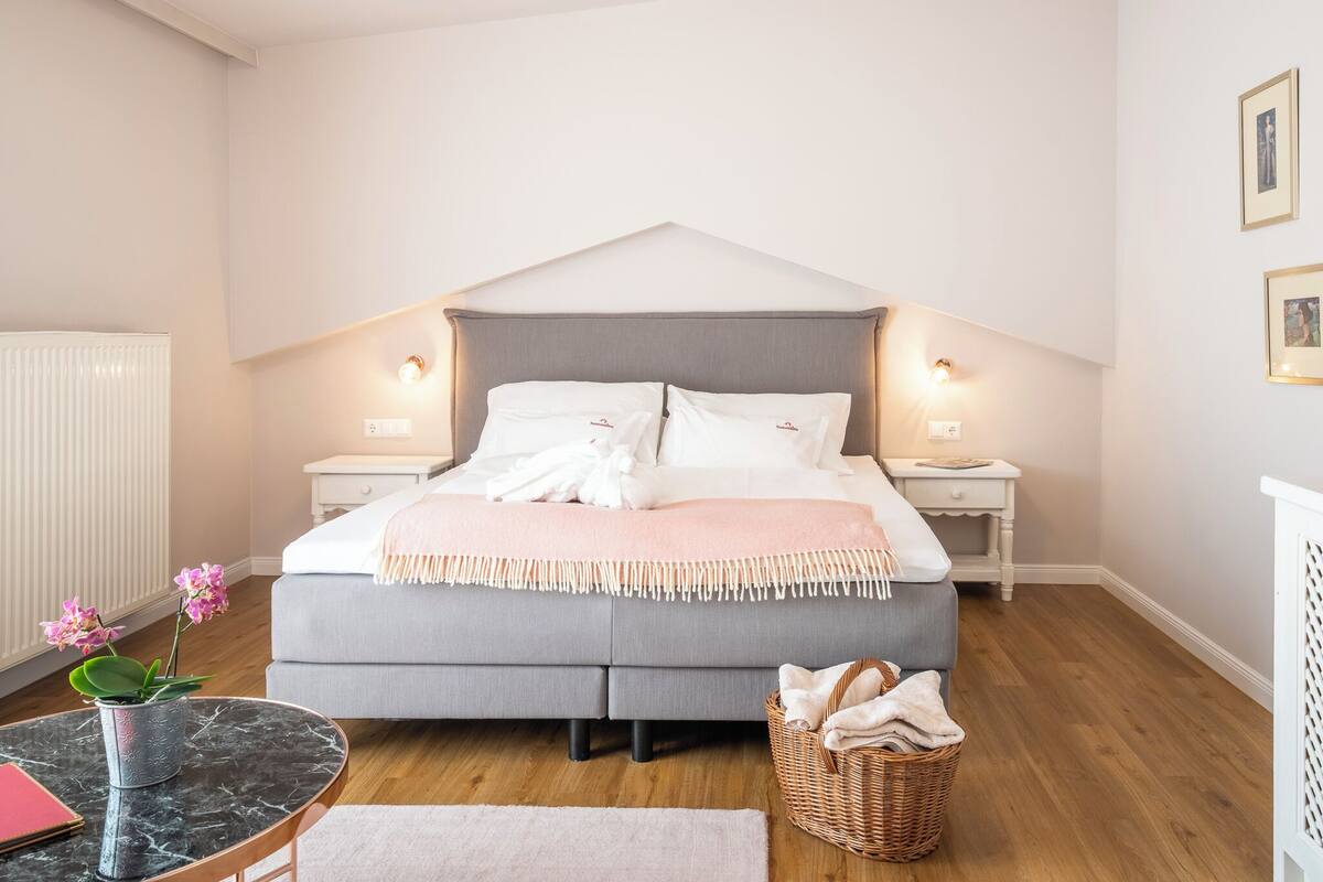 A bedroom with a bed, bedside tables, a nightstand, a small table with a vase, a wicker basket, and a radiator.