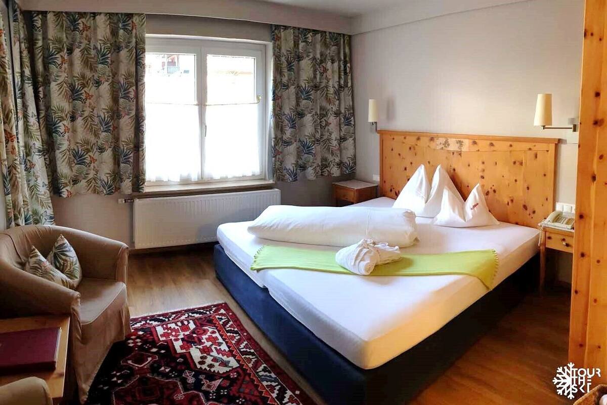 A hotel room with a bed, a chair, a window with curtains, a bedside table, and a lamp.