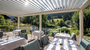 Un área de comedor al aire libre con mesas preparadas para dos, cubierta por una pérgola blanca, con vistas a una piscina y abundante vegetación.