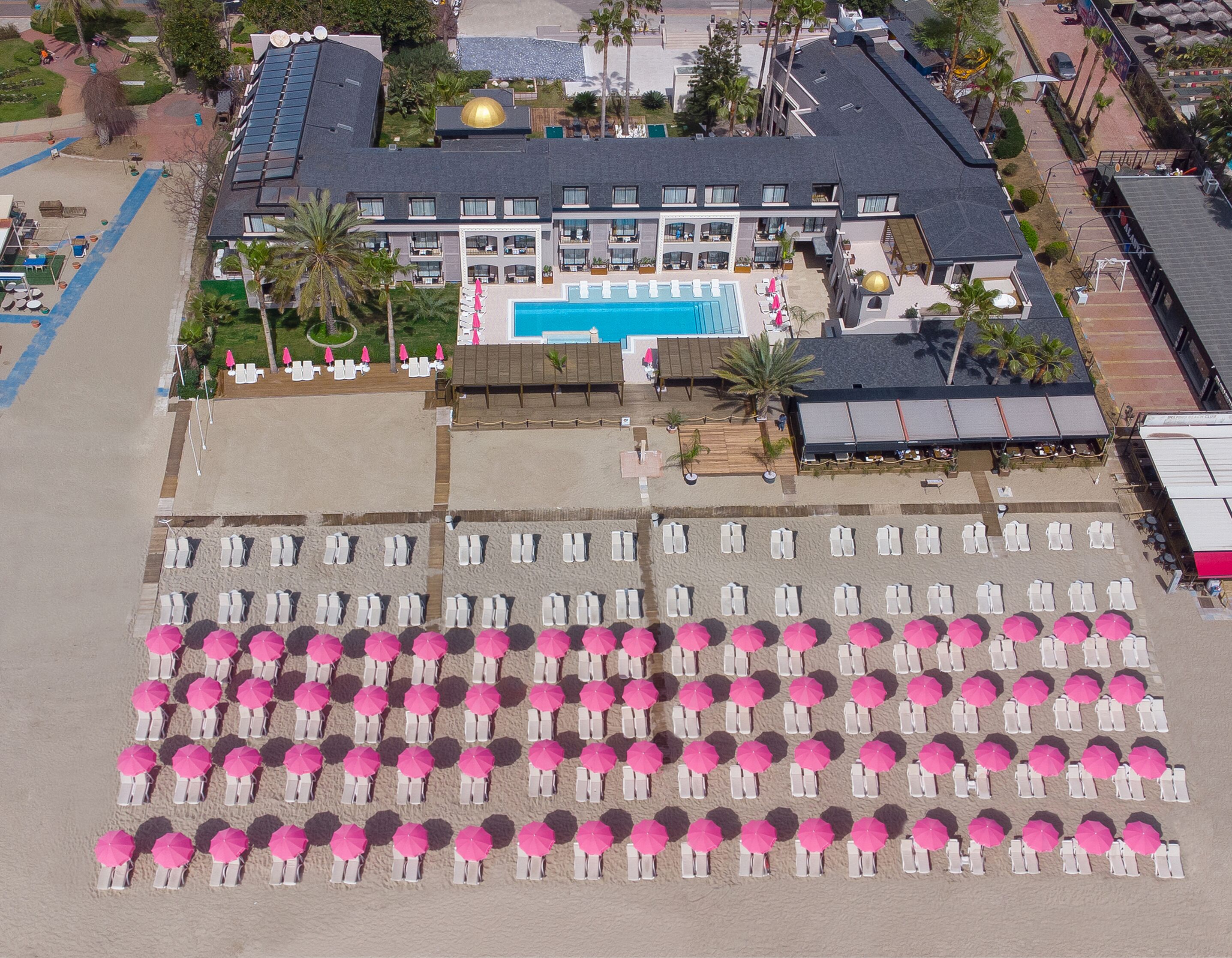 An aerial view of a hotel with a swimming pool, multiple beach umbrellas, and sun loungers arranged in rows.