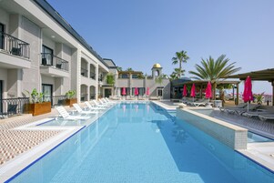 Una piscina con sillones de descanso y sombrillas, rodeada por un edificio con balcones y palmeras.