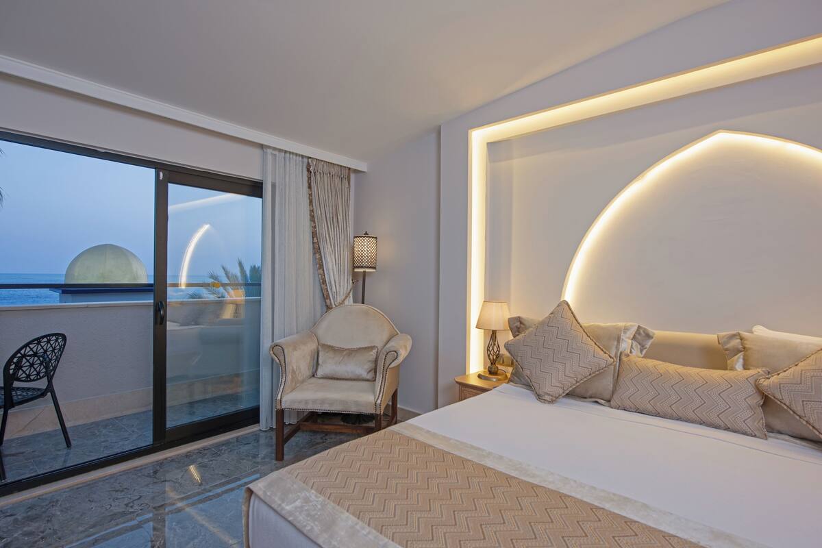 Ein Hotelzimmer mit Bett, Stuhl, Nachttisch, Lampe und Blick auf das Meer durch eine Schiebetür.
