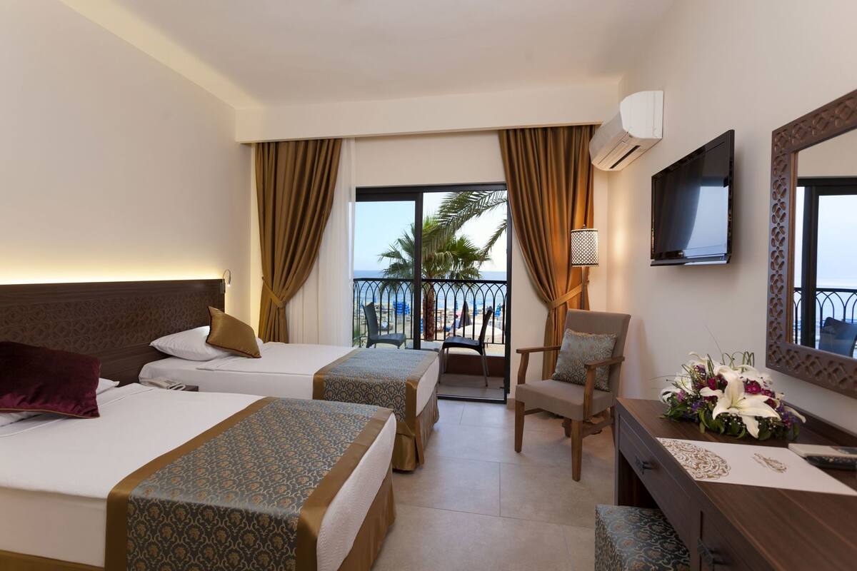 Ein Hotelzimmer mit zwei Betten, einem Schreibtisch, einem Sessel, einem Fernseher und einem Balkon mit Blick auf den Strand.
