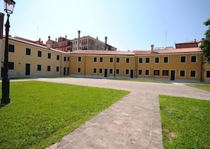 Une cour avec un chemin pavé et un bâtiment doté de nombreuses fenêtres et d’un toit recouvert de tuiles rouges.