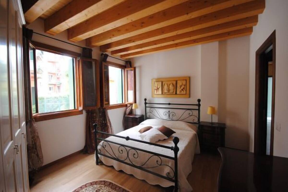 A bedroom with a wooden ceiling, a bed with a metal frame, a nightstand with a lamp, and a window with curtains.