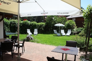 An outdoor patio area with tables and chairs, a large umbrella, and a view of a well-maintained garden. - Activ Pension Bergfrieden (Leutasch)