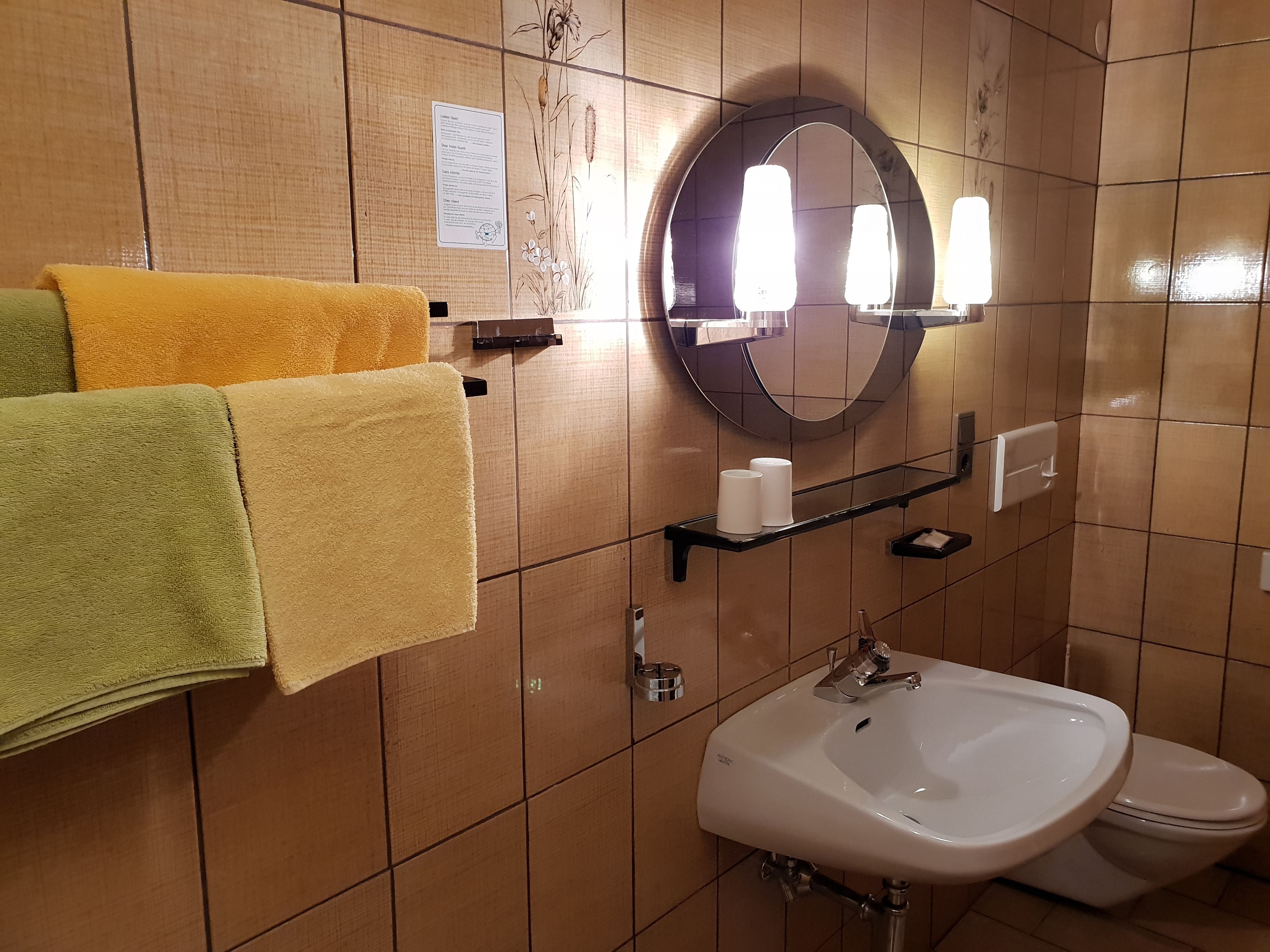 A bathroom with a sink, toilet, and a mirror with lights. There are towels hanging on a rack and a shelf with toiletries.
