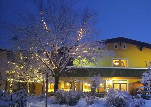 A hotel with a yellow facade, illuminated windows, and snow-covered trees. - Activ Pension Bergfrieden (Leutasch)