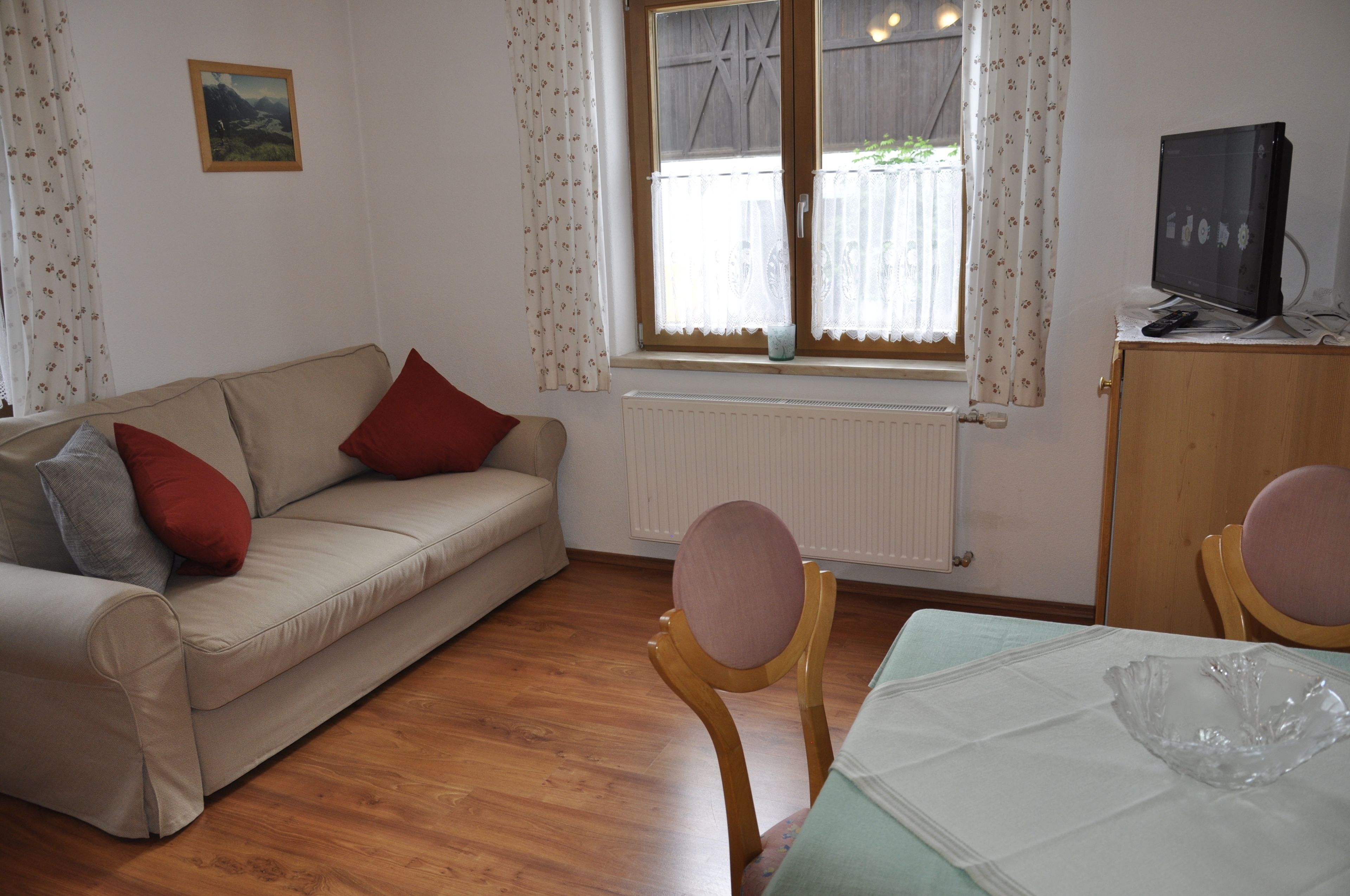 A living room with a beige sofa, red and grey cushions, a wooden chair, a small table with a white tablecloth, and a television on a wooden cabinet.