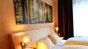 Una habitación de hotel con cabecera de madera, dos camas, una mesita de noche con una lámpara y un cuadro de un bosque en la pared.