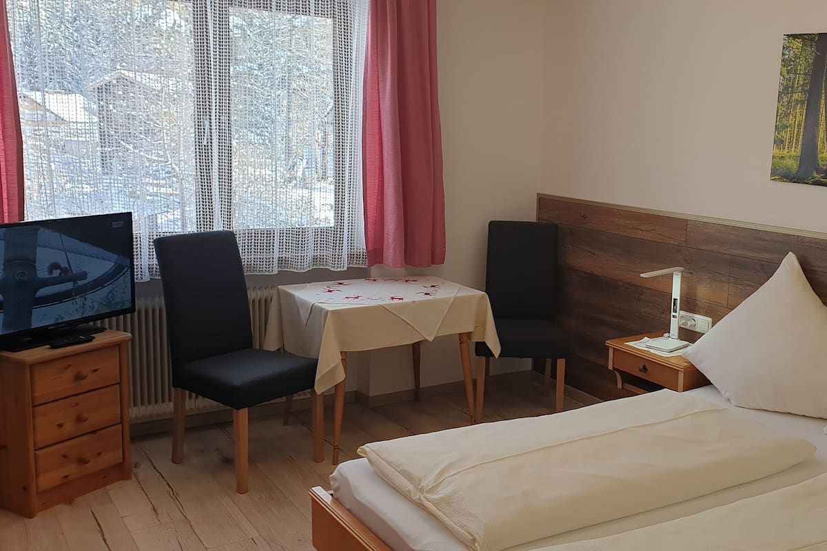 Una habitación de hotel con una cama, un escritorio con televisor, una mesa con mantel floreado y un cuadro de un bosque en la pared.