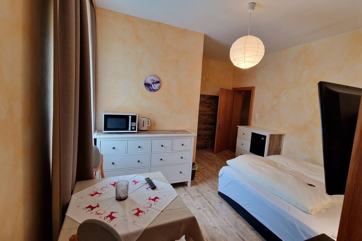 Una habitación pequeña y compacta con una cama, una mesita de noche con una taza, un microondas, un mueble pequeño y una puerta que da a otra habitación.