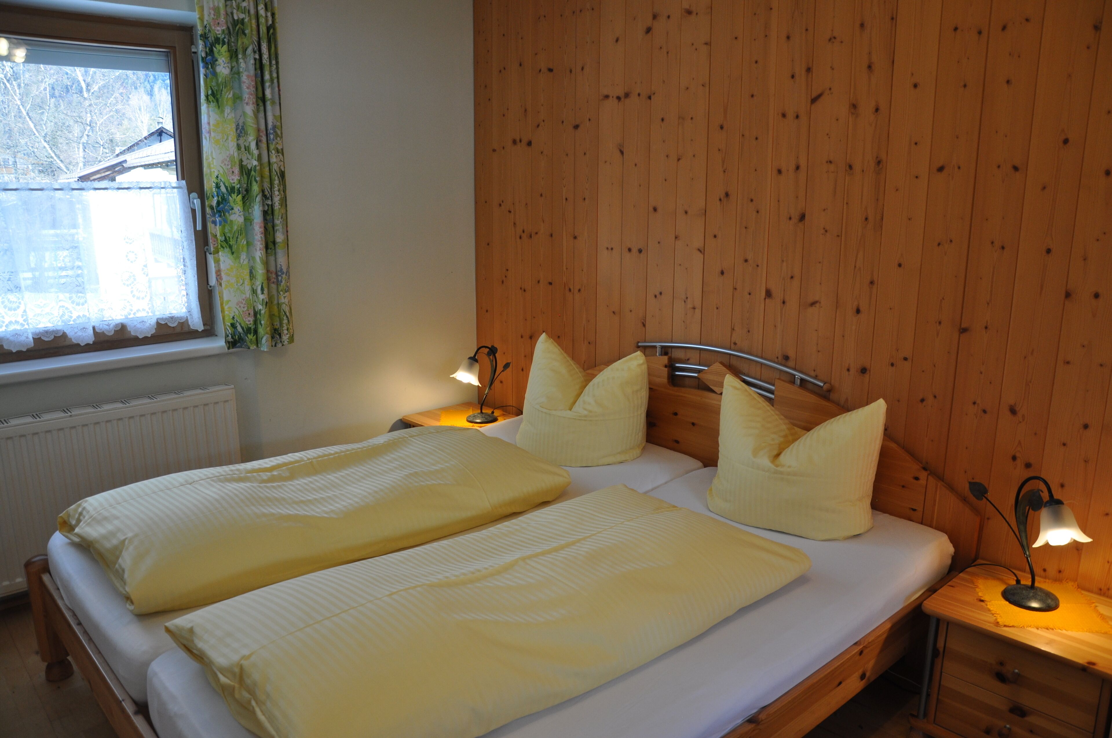 Uma cama de casal com duas almofadas, uma mesa de cabeceira com abajur e uma janela com cortinas.