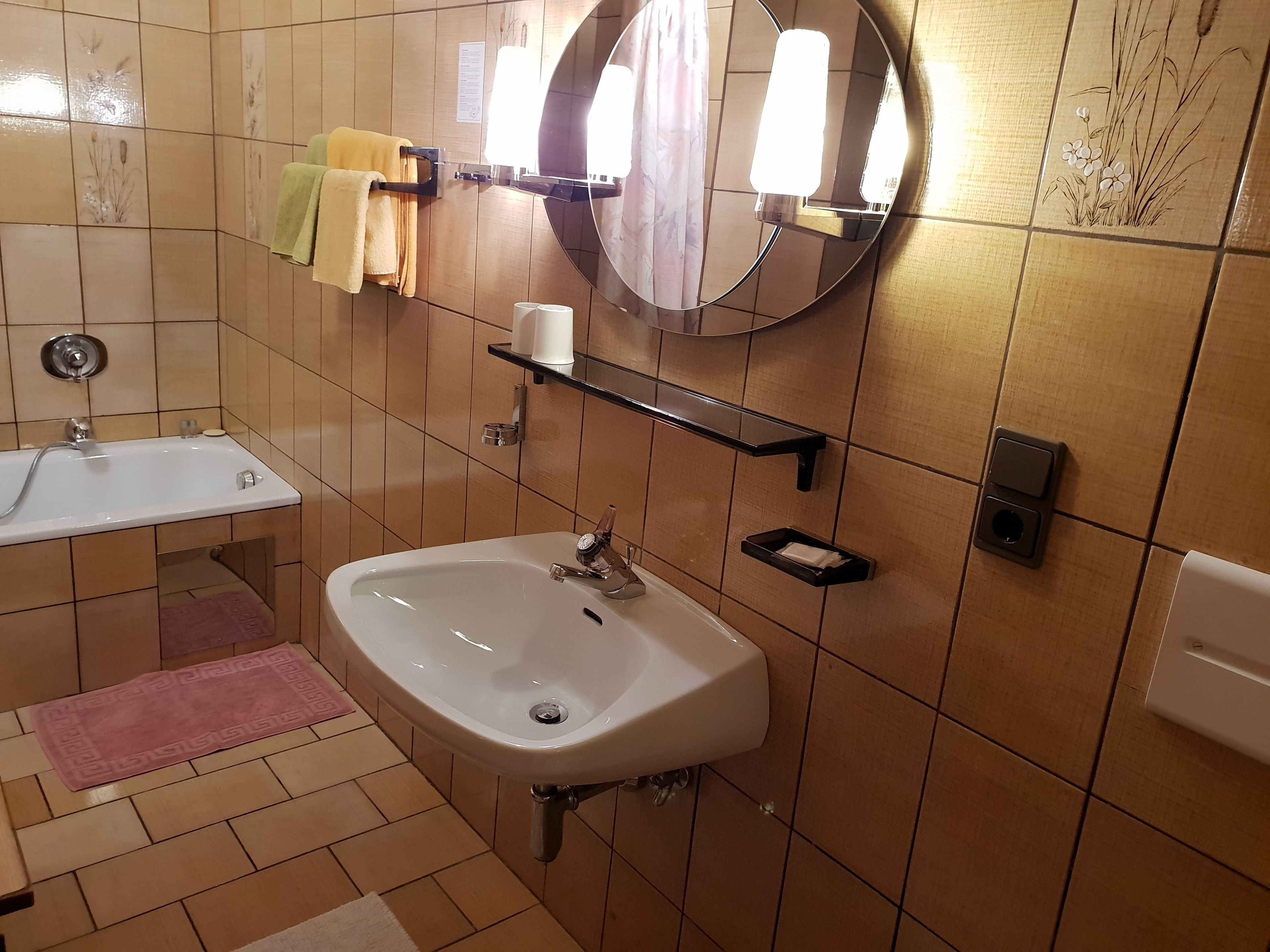 Kylpyhuone, jossa on pesuallas, peili, pyyhekoukku ja hylly, jossa on hygieniatuotteita.