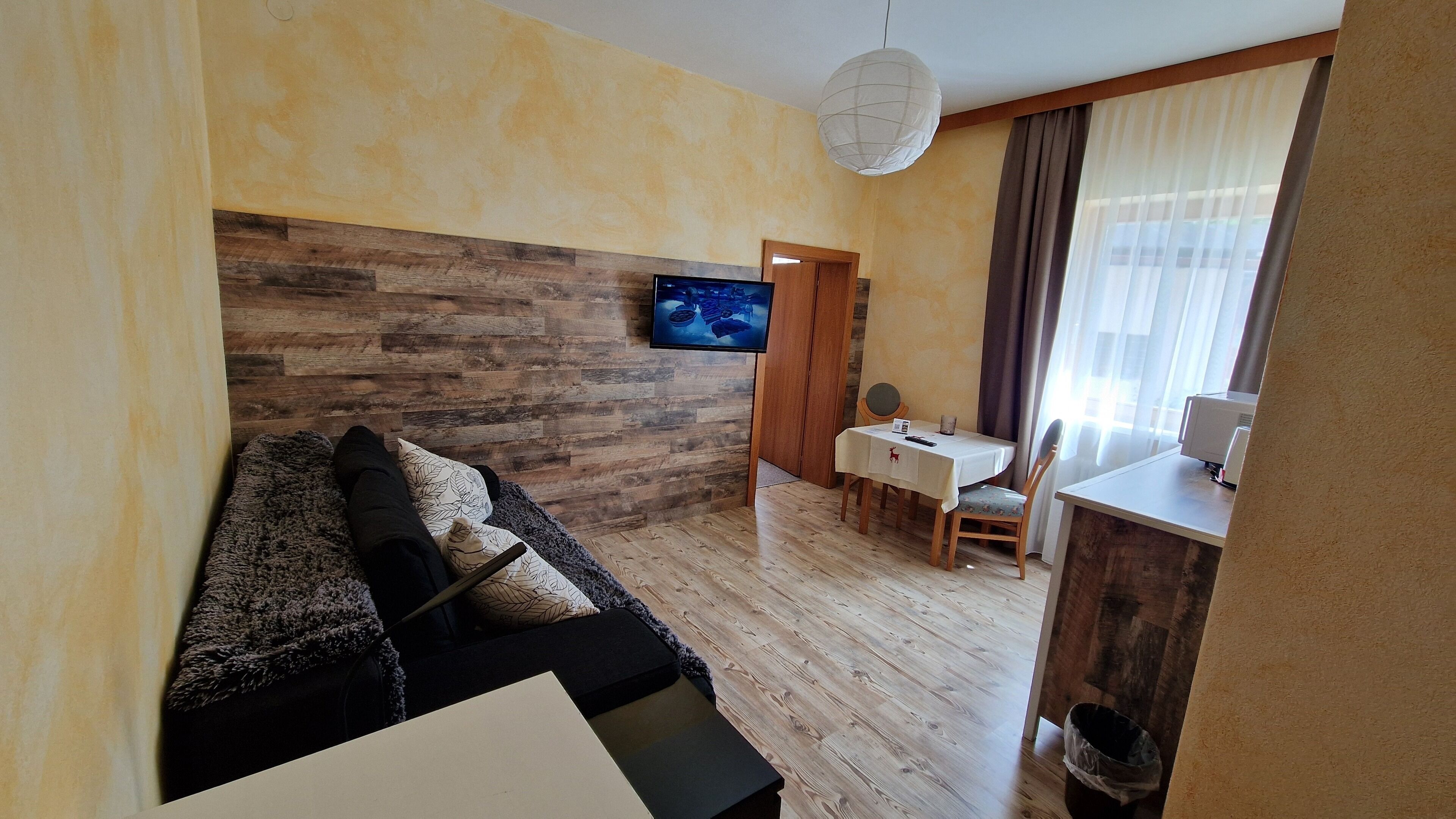 Een woonkamer met een houten wand, een bank, een televisie, een eettafel met stoelen en een keukenhoek.