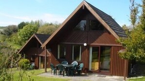 Eine Holzhütte mit geneigtem Dach, großen Fenstern und einer Terrasse mit Sitzgelegenheit im Freien.