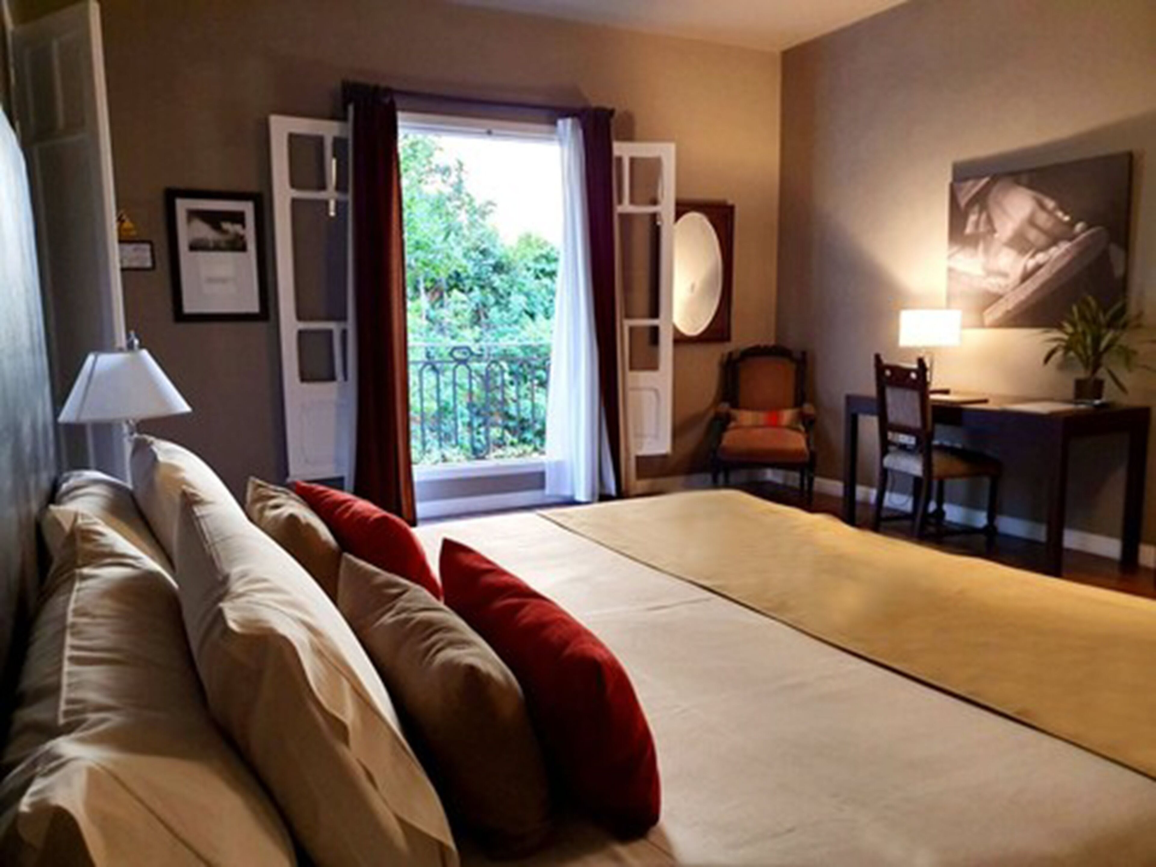 A hotel room with a bed, a desk, a chair, a lamp, and a view of greenery through an open door.