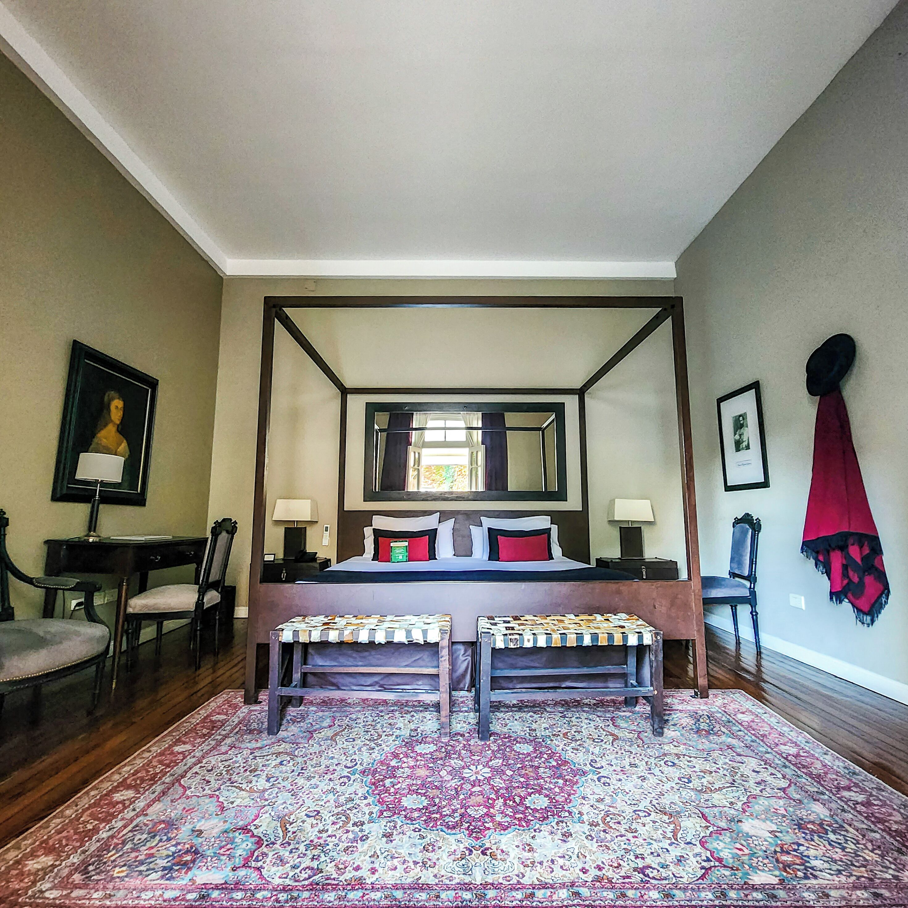 A four-poster bed with a mirror above, a desk with a chair, a bench, and a patterned rug.