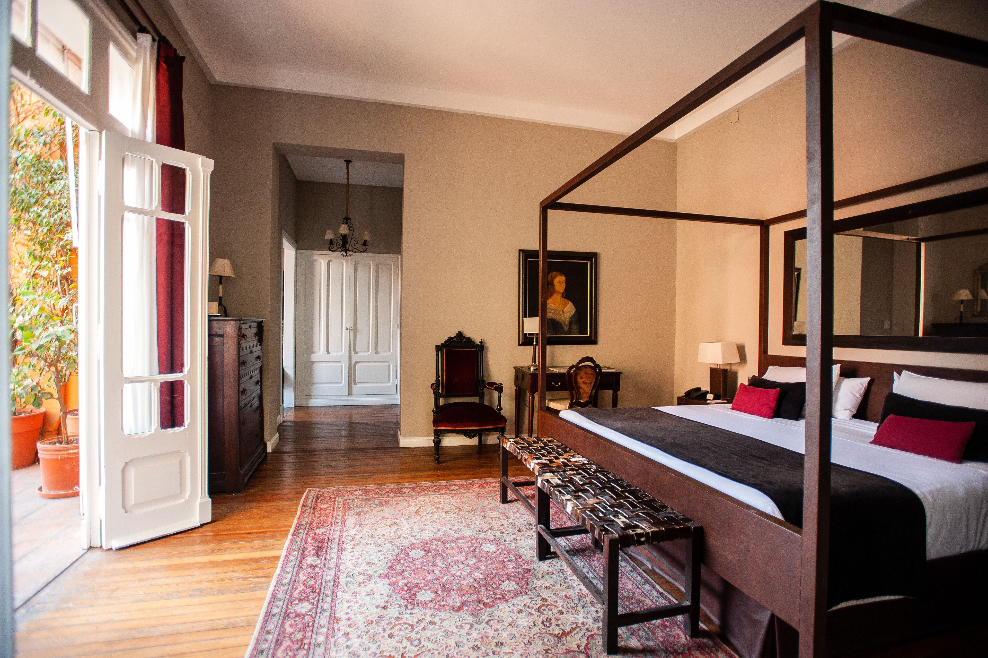 A four-poster bed with a bench, a chair, a desk, and a painting in a room with wooden floors and a large window.