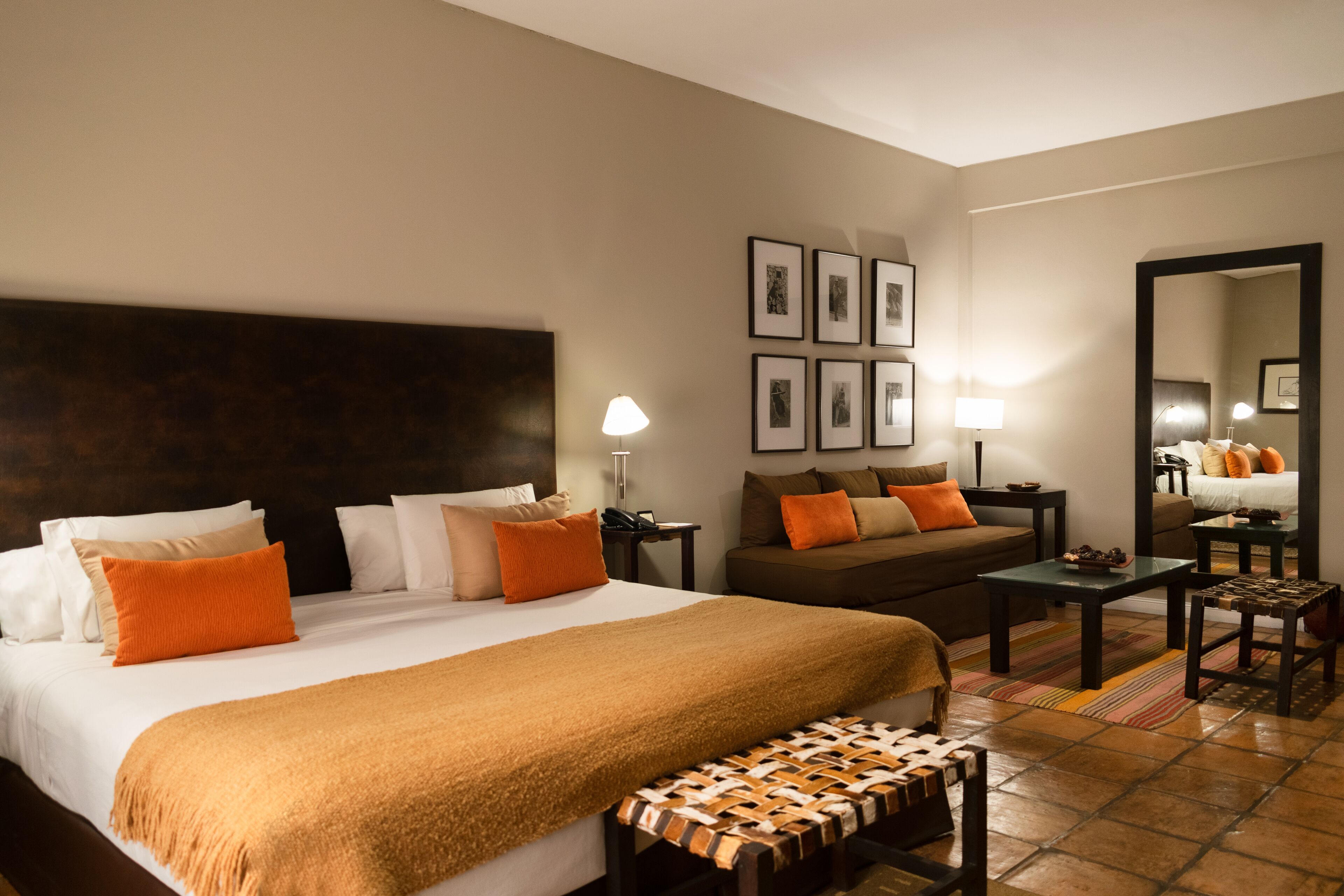 A hotel room with a large bed, a sofa, a small table, a bench, a lamp, and a mirror.